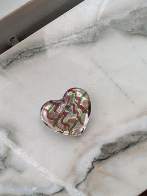 Hjärta i glas med mönster - Unikt hjärta i glas med vågigt mönster i silver, rött och grönt på framsidan och skimrande rosa baksida. Perfekt som dekoration eller smyckesdel. Hålet i mitten gör det enkelt att trä på ett band eller kedja.