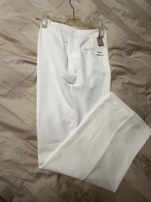 Vita kostymbyxor från H&M - Säljer ett par stilrena vita kostymbyxor från H&M. Byxorna har raka ben, hög midja och pressveck framtill. Klassisk design med fickor i sidan och hällor för skärp. Perfekta för en clean och fräsch look. Materialet är mjukt och har en lätt stretch.
