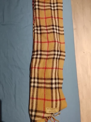 Rutig halsduk från Burberry i cashmere - Klassisk halsduk från Burberry i 100% cashmere med det ikoniska rutmönstret i beige, svart, vitt och rött. Halsduken har fransar i ändarna och är tillverkad i England. Perfekt accessoar för att lyfta din outfit.