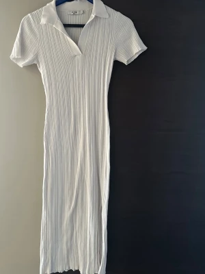 Vit ribbad klänning från NA-KD - Säljer en vit ribbad klänning från NA-KD med krage och v-ringning. Klänningen har korta ärmar och är figurnära med en lång siluett. Perfekt för dig som gillar stilrena och enkla plagg med en twist.
