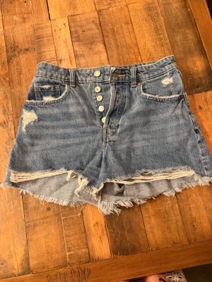 Blåa högmidjade jeansshorts med knappar - Säljer ett par blåa högmidjade jeansshorts med fransig kant och slitna detaljer. Shortsen har fem knappar framtill, klassiska fickor och råa kanter för en avslappnad look. Perfekta för sommaren och ger en cool vintage vibe.