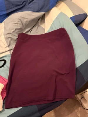 Mörklila kortkjol i stretchigt tyg - En enkel och stilren mörklila kortkjol i stretchigt material. Kjolen har en rak passform och är perfekt att matcha med både t-shirt och blus. Inga synliga detaljer eller mönster, bara clean och snygg.