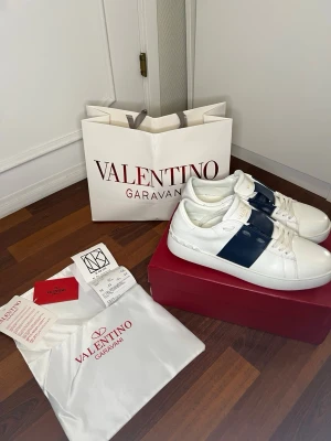 Valentino Garavani Open sneakers vit/blå - Snygga Valentino Garavani Open sneakers i vitt skinn med bred mörkblå rand över sidan. Klassisk rund tå, vita snören och diskret logga på plösen. Kommer med originalpåse och box. Fråga om de e något du undrar över. Dom har tecken på användning men det är inget man tänker på. Fråga gärna om fler bilder.
