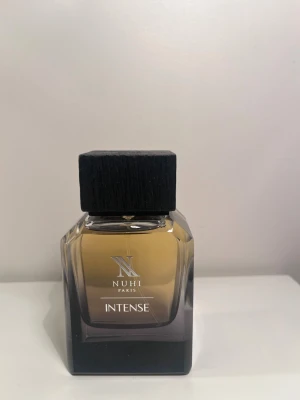 Nuhi Paris Intense parfym - Nuhi Paris Intense herrparfym i en stilren, fyrkantig glasflaska med mörk toning nedtill och svart trälock. Flaskan har en elegant design med tydlig logga och text på framsidan. Perfekt för dig som gillar exklusiva dofter och snygg inredningsdetalj på hyllan. Endast använd få gånger se bild