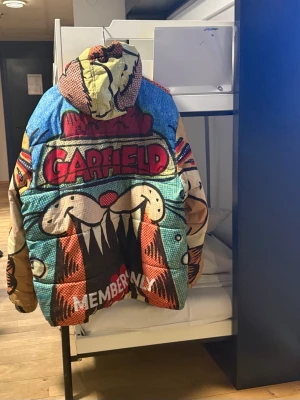 Färgglad Garfield teddyjacka Members Only - Unik teddyjacka med Garfield-motiv från Members Only. Jackan har färgglada serietidningsinspirerade tryck i rött, blått, beige och svart. Utrustad med huva och dragkedja framtill. Perfekt för dig som vill sticka ut med en oversized och mysig look.