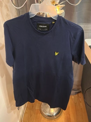 Mörkblå t-shirt från Lyle & Scott - Snygg mörkblå t-shirt från Lyle & Scott med klassisk passform och gul broderad logga på bröstet. Tillverkad i mjuk bomull och har rund halsringning samt korta ärmar. Perfekt för en stilren och enkel look.
