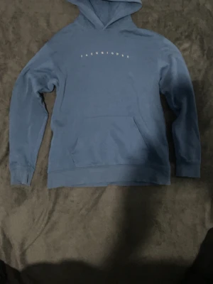 Blå hoodie från Jack & Jones - Snygg blå hoodie från Jack & Jones med vit text på bröstet. Klassisk modell med huva och stor magficka framtill. Perfekt för en avslappnad och trendig stil. Mjuk bomullskänsla och ribbade muddar vid ärmslut och nederkant.