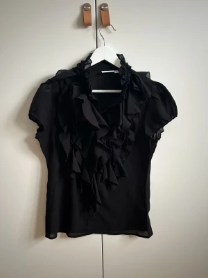 Svart volangblus med kort ärm från Saint Tropez - Stilren svart blus med korta ärmar och volangdetaljer framtill från Saint Tropez. Blusen har volanger och är tillverkad i ett tunt, lätt material som ger ett elegant fall. Perfekt för dig som gillar en chic och trendig look. Skickar snabbt!