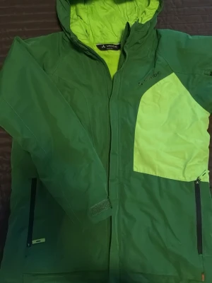Grön vinterjacka från Vaude stl 158/164 - Grön vinterjacka från Vaude till ungdom med limegula detaljer och huva. Jackan har dragkedja framtill, flera fickor och justerbara ärmslut med kardborre. Perfekt för utomhusaktiviteter och träning i kallare väder. Snölås och plats för liftkort. Tillhörande är något mer använda thermobyxor och medföljer. 
