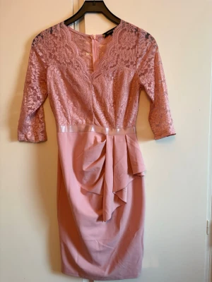 Rosa spetsklänning med volangdetalj - Söt rosa klänning med spets upptill och trekvartsärmar. Kjolen har en snygg volangdetalj framtill och markerad midja med satinband. Klänningen har dragkedja i ryggen och är figurnära i modellen. Har bara använt den en gång. Så riktigt nyskick!