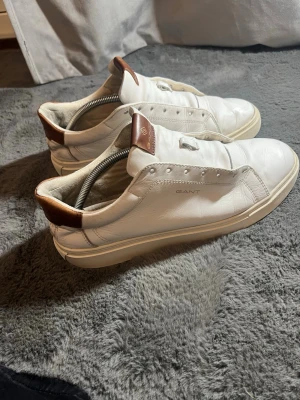 Vita sneakers från Gant i skinn - Stilrena vita sneakers från Gant i skinn med bruna detaljer på häl och plös. Klassisk låg modell med rund tå och platt sula. Diskret Gant-logga på sidan och snygga sömmar för en clean look. Saknar snören 
