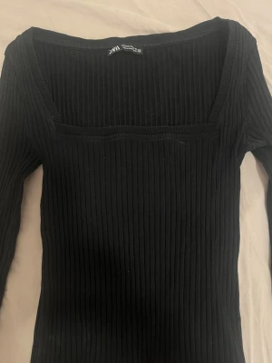 Svart ribbad topp från Zara - Säljer en svart ribbad topp från Zara med lång ärm och fyrkantig ringning. Toppen är i stretchigt material och har en slim passform som sitter snyggt på kroppen. Perfekt att styla med jeans eller kjol.