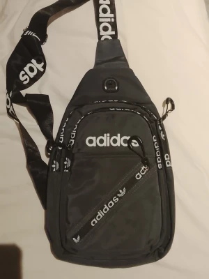 Svart Adidas handväska med logga - Snygg svart handväska från Adidas med vit logga och bred axelrem med Adidas-tryck. Väskan har flera fack med dragkedja och en sportig design. Perfekt för dig som vill ha en trendig och praktisk väska till vardagen.