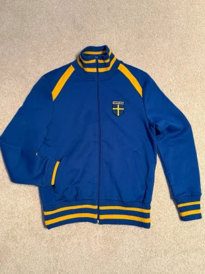 Blå-gul Jack&Jones Sverige retro track jacket - Snygg blå retro track jacket med gula detaljer och ränder. Dragkedja framtill, ståkrage med ränder och broderad Sverige-logga på bröstet. På ryggen står det SWEDEN i gult. Perfekt inför OS!
