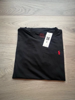 Svart t-shirt från Polo Ralph Lauren - Svart t-shirt från Polo Ralph Lauren med klassisk passform och korta ärmar. T-shirten har en röd broderad logga på bröstet och är tillverkad i mjuk bomull. Perfekt för en stilren och enkel look.
