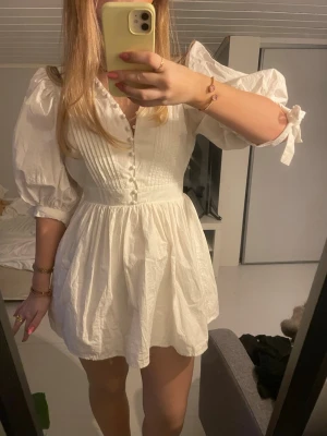 Vit kort klänning med puffärm - Supersöt vit kort klänning med puffärmar och knappar framtill. Klänningen har v-ringning, markerad midja och fina veckdetaljer som ger en romantisk känsla. Tillverkad i ett lätt bomullstyg, perfekt för varmare dagar.💞