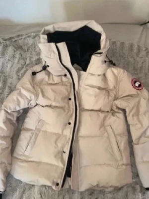 Beige dunjacka från Canada Goose - Snygg beige dunjacka från Canada Goose med huva och svart innerfoder. Jackan har dragkedja och knappar framtill, samt den klassiska loggan på ärmen. Perfekt för kalla dagar och har en puffig, quiltad design.