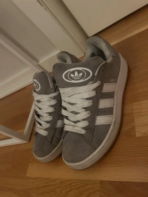 Grå Adidas Campus sneakers mocka - Säljer ett par grå Adidas Campus sneakers i mocka med vita detaljer och klassiska tre ränder på sidan. Skorna har rund tå, platt sula och snörning. Adidas-logga på plösen och sidan. Perfekta för dig som gillar stilrena och tidlösa sneakers. Tvättas innan de skickas då de mesta på skon är ytligt smuts 