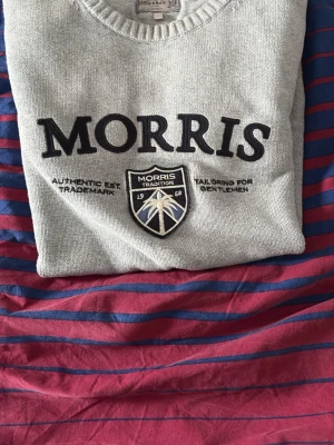 Grå stickad tröja från Morris - Snygg grå stickad tröja från Morris med broderad logga och emblem framtill. Rund halsringning och ribbade muddar. Perfekt för en stilren och klassisk look.