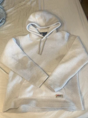 Vit fleecehoodie från Everest - Mysig vit hoodie från Everest i fluffigt fleecematerial. Hoodien har en stor huva med dragsko och en praktisk magficka framtill. Perfekt för dig som gillar en avslappnad och varm stil. Everest-logga med bergmotiv på fickan.