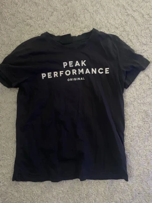 Mörkblå Peak Performance t-shirt - Snygg mörkblå t-shirt från Peak Performance med vit text på bröstet. Klassisk rund halsringning och korta ärmar. Tillverkad i mjuk bomull som känns skön mot huden. Perfekt för en avslappnad och sportig stil.