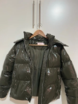 Grön jacka från Nelly - Snygg mörkgrön pufferjacka från Nelly med glansig finish och quiltad design. Jackan har huva, dragkedja framtill och en loose passform som ger en trendig look. Perfekt för kalla dagar när du vill vara både varm och stylish.