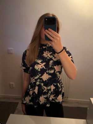 Blommig blus med öppen rygg - Mörkblå blus med ljusa blommor och peplumvolang. Blusen har korta ärmar och en snygg öppen rygg med knytband upptill. 