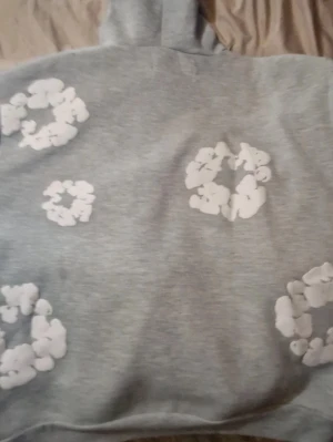 Grå hoodie med vita blommor - Snygg grå hoodie med huva och stora vita blommönster tryckta över hela tröjan. Hoodien har en avslappnad passform och är tillverkad i mjukt material, perfekt för en chill och trendig look. Denna hoodie är i storlek S