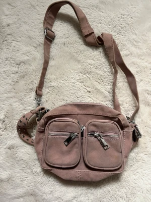Noella väska - Snygg rosa axelväska i mocka med två framfickor med dragkedja och justerbart axelband med silverfärgade nitar. Perfekt för att bära det viktigaste och ger en cool touch till din outfit. I den lilla storleken