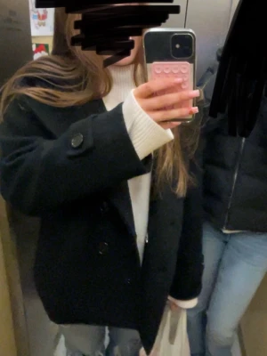 Svart Design by Si kappa - Stilren svart kappa med bred krage och två stora knappar framtill. Modellen är något kortare och har en rak passform med långa ärmar och dekorativa knappar vid ärmsluten. Perfekt för dig som gillar en klassisk och enkel look.