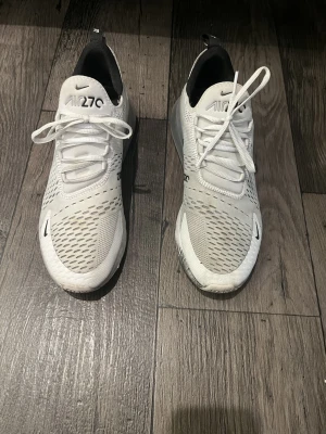 Nike Air Max 270 - Nike Air Max 270 sneakers i vitt med svarta detaljer och stor synlig luftbubbla i hälen. Ovandelen är i mesh för bra ventilation och har klassisk snörning. Snygg och sportig design med logga på sidan och plös.