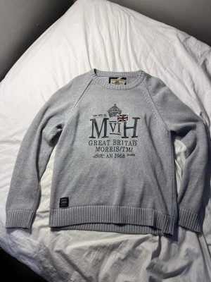 Ljusgrå stickad tröja Morris - Ljusgrå stickad tröja från Morris med tryck framtill som inkluderar brittisk flagga och text. Tröjan har rund hals, ribbade muddar och nederkant. Perfekt för dig som gillar klassisk och stilren design med en sportig touch.