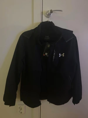 Svart vindjacka från Under Armour - Svart vindjacka från Under Armour med klassisk passform och dragkedja framtill. Jackan har lång ärm, diskret logga på bröstet och är tillverkad i lätt syntetmaterial som passar perfekt för blåsiga dagar.
