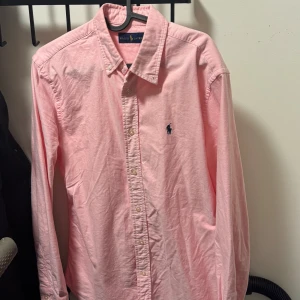 Rosa skjorta från Ralph Lauren S - Snygg rosa skjorta från Ralph Lauren i storlek S. Klassisk button-down krage, långärmad och med det ikoniska blå broderade logot på bröstet. Skjortan är tillverkad i mjuk bomull och har knappar framtill. Perfekt för dig som gillar stilrena och tidlösa plagg.