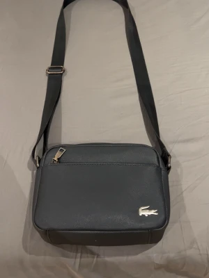 Svart axelväska från Lacoste - Snygg svart axelväska från Lacoste med justerbar rem och silverfärgad dragkedja. Väskan har en stilren design med Lacostes ikoniska krokodillogga i metall på framsidan. Perfekt storlek för det viktigaste och tillverkad i slitstarkt syntetmaterial/ läder.