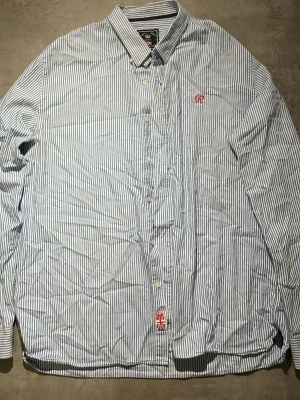 Blårandig skjorta Robbie Tailor XL - Snygg blårandig skjorta från Robbie Tailor i slim fit-modell. Skjortan har klassisk krage, knappar framtill och en liten röd broderad logga på bröstet. Tillverkad i bomull och passar perfekt till en clean och stilren look.