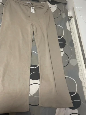 Beige mjukisbyxor från 157 - Snygga beige mjukisbyxor från 157 med breda ben och dragsko i midjan. Byxorna har en avslappnad passform och är gjorda i mjukt bomullsmaterial, perfekta för en chill och bekväm stil.