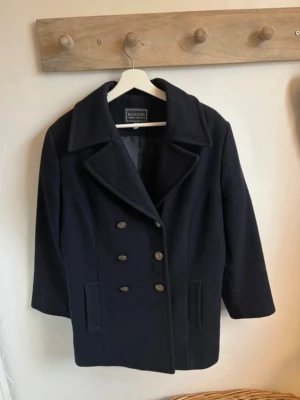 Marinblå skepparkavaj från Havana - Stilren marinblå skepparkavaj från Havana med klassisk dubbelknäppning i guldtonade knappar och bred krage. Jackan har raka fickor framtill och är tillverkad i ett ulligt material som ger en tidlös look.