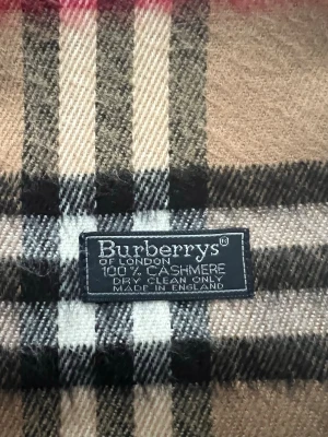 Beige rutig halsduk från Burberrys - Klassisk beige halsduk från Burberrys i 100% kashmir. Den har det ikoniska rutmönstret i beige, svart, vitt och rött samt fransar i båda ändar. Perfekt accessoar för att lyfta din outfit med en tidlös brittisk stil.