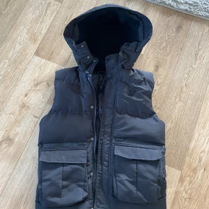 Svart dunväst med huva från SikSilk - Svart dunväst från SikSilk med stor huva och flera praktiska fickor framtill. Västen har dragkedja och tryckknappar, samt justerbar dragsko i huvan. Perfekt för lager-på-lager och riktigt snygg till streetwear-stilen.