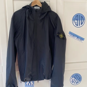 Svart vindjacka från Stone Island (Helt ny) - Säljer en svart vindjacka från Stone Island med huva och dragkedja framtill. Jackan har den klassiska loggan på vänster ärm och är tillverkad i borstad bomull med vattenavvisande finish.Jackan sitter som en stor M men lutar lite mer åt L hållet. men  Perfekt för dig som gillar stilrena och funktionella plagg. 