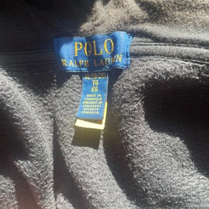 Svart hoodie från Polo Ralph Lauren XL - Svart hoodie från Polo Ralph Lauren i storlek XL, i barn storlek(170 cm). Klassisk design med dragkedja framtill, huva och två fickor. Liten röd logga broderad på bröstet. Mjuk och skön bomullsblandning, perfekt för chill dagar. 