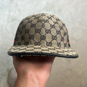 Gucci keps med GG-mönster och röd detalj - Snygg beige keps från Gucci med klassiskt GG-mönster i svart över hela kepsen. På sidorna finns en bred röd och svart rand som ger extra lyxig känsla. Justerbar rem bak i svart skinn. Perfekt för dig som vill sticka ut med designerstil.