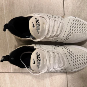 Nike Air Max 270 vita sneakers - Nike Air Max 270 sneakers i vitt med svarta detaljer och stor synlig Air-enhet i hälen. Ovandelen är i mesh med ventilerande mönster och snörning framtill. Snygg och sportig look med svart Nike-logga på sidan och AIR-tryck bak. Perfekta för dig som gillar streetstyle. Väldigt bra skick, nypris 1500