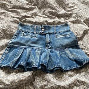 Blå jeanskjol med volangkant - Supersöt blå jeanskjol med volangkant och markerad midja. Kjolen har tre knappar framtill, bälteshällor och fejkade fickor. Perfekt för dig som gillar en cool och lekfull stil. Klassisk denim-look med slitningar och plisserad nederdel.