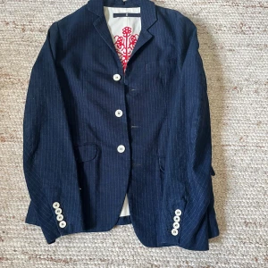 Mörkblå randig kavaj från Dondup - Snygg mörkblå kavaj från Dondup med tunna vita ränder och vita knappar fram och vid ärmslut. Insidan har ett unikt rött tryck med Dondup-logga. Kavajen har klassisk krage, två fickor fram och är tillverkad i bomull. Perfekt för dig som gillar stilrena detaljer.