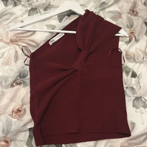 Vinröd oneshoulder topp från Zara - Supersnygg vinröd oneshoulder topp från Zara med ribbad struktur och knyt-detalj framtill. Toppen är ärmlös och har en cool asymmetrisk form som ger en trendig vibe. Perfekt att styla med jeans eller kjol för en chic look.