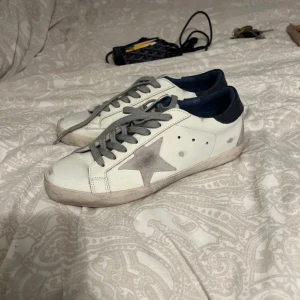 Golden Goose Superstar sneaker - Golden Goose Superstar sneakers i vitt skinn med grå stjärndetalj på sidan, grå snörning och mörkblå häl. Klassisk låg modell med platt sula och rund tå. Snyggt slitna detaljer för en cool, avslappnad vibe.