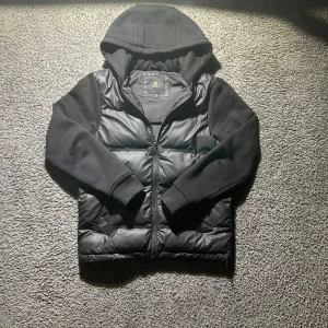 Svart cardigan från next - Svart cardigan från Next i storlek XS. Jackan har quiltad kropp i syntetmaterial, mjuka ärmar och huva i sweatshirt-tyg. Stängs med dragkedja framtill och har ribbade muddar vid ärmslut. Perfekt för kyliga dagar. Ny pris 1300 använd fåtal gånger