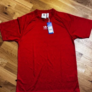 Röd Adidas t-shirt med mönster XS - Snygg röd t-shirt från Adidas med diskret geometriskt mönster och klassisk vit Adidas-logga på bröstet. Modellen är kortärmad och har rund hals. Materialet är mjuk bomull som känns skönt mot huden. Perfekt för dig som gillar sportig och stilren streetwear.